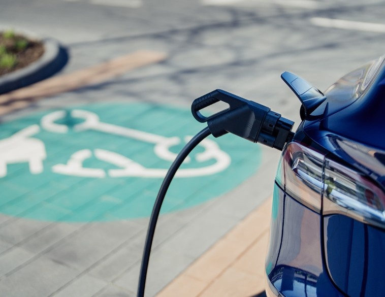 Gros plan sur l'arrière d'un véhicule électrique bleu en train de se recharger, avec un câble branché dans la prise et un symbole de stationnement pour voiture électrique peint en vert sur le sol en arrière-plan.