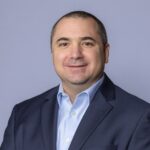 Portrait professionnel de Phil Diogenes, le nouveau vice-président, Développement des affaires chez OK Tire. Il porte un veston bleu marine foncé et une chemise boutonnée bleu clair sur un fond neutre et uni.
