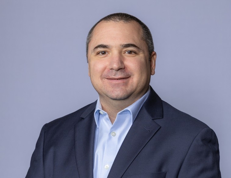 Portrait professionnel de Phil Diogenes, le nouveau vice-président, Développement des affaires chez OK Tire. Il porte un veston bleu marine foncé et une chemise boutonnée bleu clair sur un fond neutre et uni.