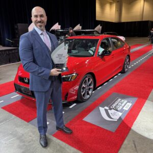 Un homme en complet bleu tenant le trophée de la Voiture canadienne de l'année de l'AJAC , debout à côté d’une Honda Civic 2026 rouge sur un tapis rouge lors du Salon international de l’auto du Canada 2026. 