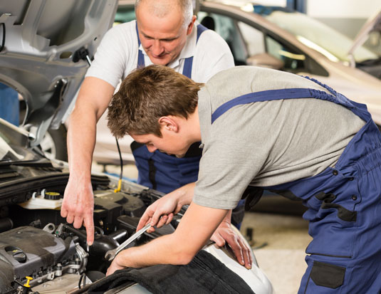 Two automotive technicians wearing blue overalls work together on a vehicle's engine in a repair shop; one man points toward a component while the other holds a wrench. 
