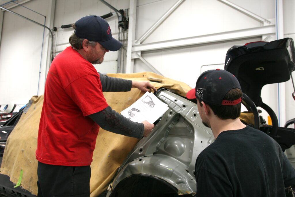 Un technicien senior vêtu d'un t-shirt rouge et d'une casquette bleue montre un document technique à un apprenti tout en examinant la structure de la carrosserie d'un véhicule dans un atelier de réparation.