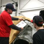 Un technicien senior vêtu d'un t-shirt rouge et d'une casquette bleue montre un document technique à un apprenti tout en examinant la structure de la carrosserie d'un véhicule dans un atelier de réparation.