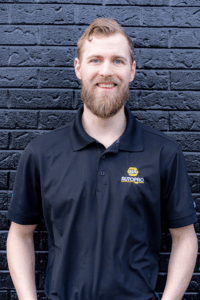 Un portrait d'Andrew Ackers, propriétaire de NAPA AUTOPRO Upper James, vêtu d’un polo noir à l’effigie de l’entreprise devant un mur de briques noires.
