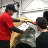 A senior technician in a red shirt and blue cap holds a technical document while pointing to a car's body structure, mentoring a junior technician in a collision repair shop.