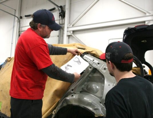 A senior technician in a red shirt and blue cap holds a technical document while pointing to a car's body structure, mentoring a junior technician in a collision repair shop.