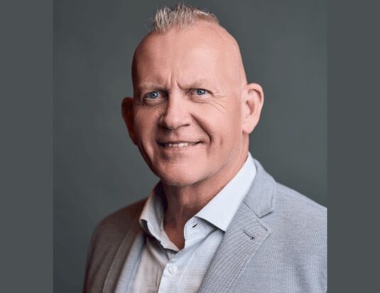 A head and shoulders professional portrait of a middle-aged Caucasian man with bright blue eyes and a short gray mohawk, smiling warmly at the camera. He wears a light gray blazer and a light blue collared shirt against a solid, neutral gray background.