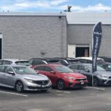 A row of pre-owned vehicles, including a silver Honda Civic and a red Kia Forte, parked in a dealership lot in front of a grey stone building. A black feather flag with the Kia logo and the words "Certified Pre-Owned" stands prominently among the cars, illustrating the current used vehicle market where dealers face challenging conditions.