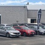 Une rangée de véhicules d'occasion, incluant une Honda Civic argentée et une Kia Forte rouge, stationnés dans la cour d'un concessionnaire devant un bâtiment gris. Un oriflamme noir avec le logo Kia et la mention « Certified Pre-Owned » est mis en évidence, illustrant les défis actuels auxquels font face les concessionnaires sur le marché de l'occasion.
