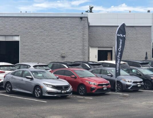 Une rangée de véhicules d'occasion, incluant une Honda Civic argentée et une Kia Forte rouge, stationnés dans la cour d'un concessionnaire devant un bâtiment gris. Un oriflamme noir avec le logo Kia et la mention « Certified Pre-Owned » est mis en évidence, illustrant les défis actuels auxquels font face les concessionnaires sur le marché de l'occasion.