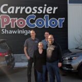 A group of four smiling people standing in front of the "Carrossier ProColor Shawinigan" sign on a black corrugated building wall, flanked by two parked vehicles.