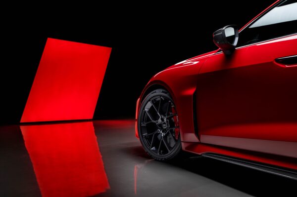 A side-angle close-up of a vibrant red Audi RS 5 in a dark studio setting, highlighting its black multi-spoke alloy wheels, red brake calipers, and Pirelli P Zero R tires. A large, glowing red light panel is reflected on the polished floor in the background.