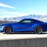 A side profile view of a vibrant blue Toyota Supra sports car parked on an asphalt road next to a concrete barrier. The background features light-brown, arid mountains under a clear blue sky with thin, wispy clouds.