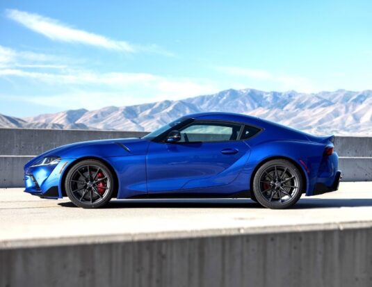 A side profile view of a vibrant blue Toyota Supra sports car parked on an asphalt road next to a concrete barrier. The background features light-brown, arid mountains under a clear blue sky with thin, wispy clouds.