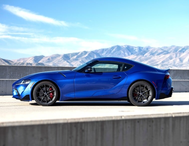 A side profile view of a vibrant blue Toyota Supra sports car parked on an asphalt road next to a concrete barrier. The background features light-brown, arid mountains under a clear blue sky with thin, wispy clouds.