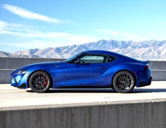 Vue de profil d'une Toyota Supra bleu vif stationnée sur une route, avec un muret en béton au premier plan et des montagnes arides en arrière-plan sous un ciel bleu clair.