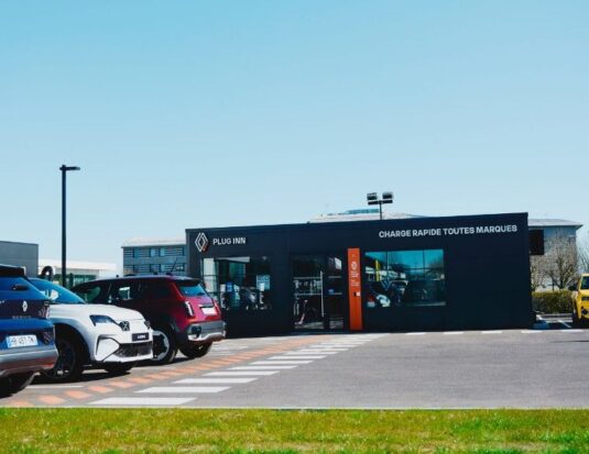 Vue extérieure d'une station de recharge Plug Inn du Groupe Renault, un bâtiment modulaire noir affichant « CHARGE RAPIDE TOUTES MARQUES ». Plusieurs voitures électriques, dont des modèles Renault et Alpine, sont garées devant.