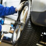 Close-up of a technician wearing blue overalls and protective gloves using a pneumatic impact wrench on a car wheel in a repair shop.