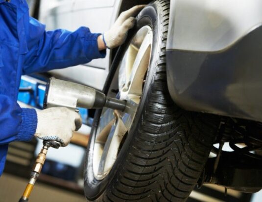 Close-up of a technician wearing blue overalls and protective gloves using a pneumatic impact wrench on a car wheel in a repair shop.