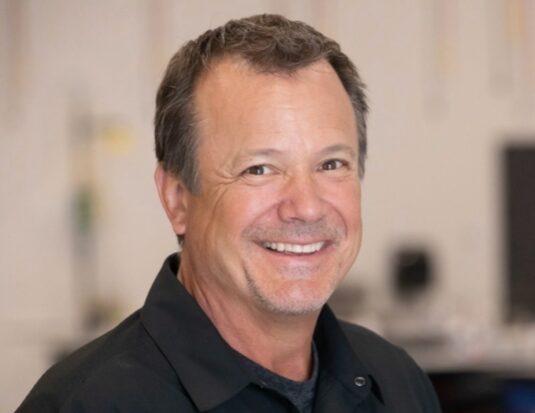 A headshot of Shawn Collins, the new Training Director for Pro Spot International, smiling and wearing a black collared shirt against a blurred background.