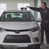A technician demonstrates an ADAS calibration tool mounted on the roof of a white Toyota Corolla in a professional collision repair facility.