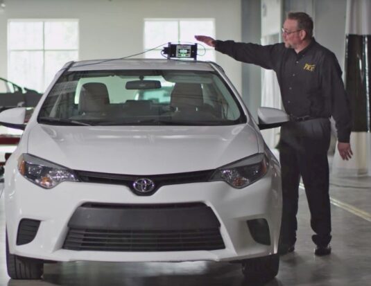A technician demonstrates an ADAS calibration tool mounted on the roof of a white Toyota Corolla in a professional collision repair facility.