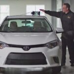 Un technicien en carrosserie effectuant un étalonnage ADAS à l'aide d'un dispositif électronique placé sur le toit d'une Toyota Corolla blanche dans un atelier de réparation professionnel