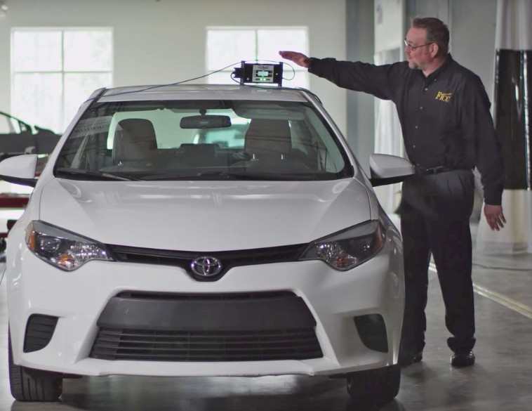 Un technicien en carrosserie effectuant un étalonnage ADAS à l'aide d'un dispositif électronique placé sur le toit d'une Toyota Corolla blanche dans un atelier de réparation professionnel