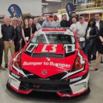 Une photo de groupe montrant une vingtaine de personnes rassemblées derrière une voiture de course rouge, blanche et noire dans un vaste atelier de carrosserie. Le véhicule est marqué des logos « Subway » et « Bumper to Bumper ».
