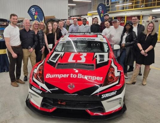 Une photo de groupe montrant une vingtaine de personnes rassemblées derrière une voiture de course rouge, blanche et noire dans un vaste atelier de carrosserie. Le véhicule est marqué des logos « Subway » et « Bumper to Bumper ».