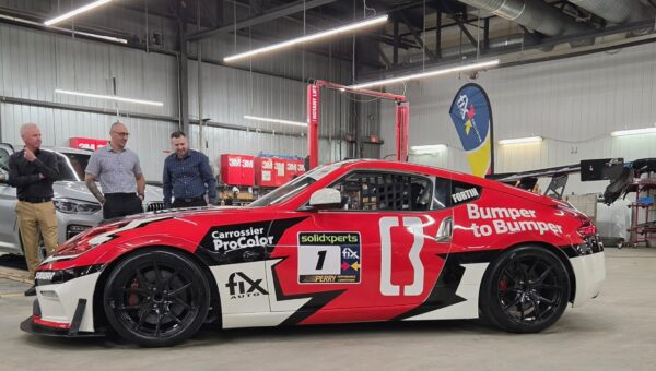 Une voiture de course rouge, noire et blanche portant le numéro 1 et le nom « Fortin » est exposée dans l'atelier de 25 000 pieds carrés de Fix Auto Lebourgneuf. Trois hommes en tenue professionnelle observent le véhicule marqué des logos de commanditaires comme « Bumper to Bumper ». 