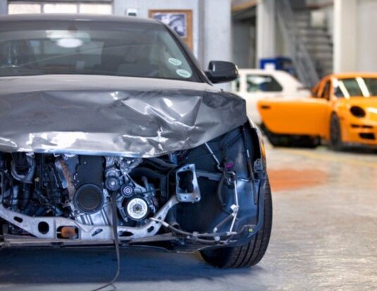 Vue avant d'une voiture grise gravement endommagée dans un atelier de carrosserie, exposant les composants internes du moteur et de la structure.