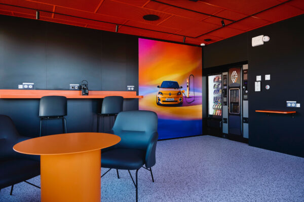 A modern indoor lounge area at a Plug Inn charging station, featuring a sleek black and orange color scheme. The space includes a round orange table with black armchairs, a long work counter with power outlets, vending machines for snacks and coffee, and a large colorful wall mural of a yellow Renault 5 electric car.