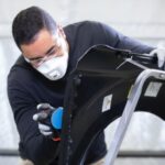 Un technicien en carrosserie portant un masque de protection et des gants, utilisant une ponceuse orbitale munie d'un disque abrasif bleu pour préparer la surface d'une aile de voiture noire sur un support.