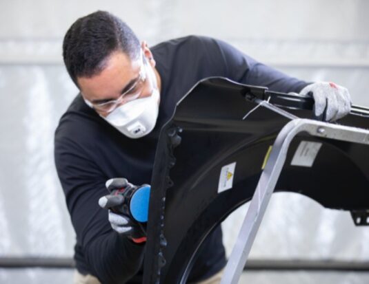 Un technicien en carrosserie portant un masque de protection et des gants, utilisant une ponceuse orbitale munie d'un disque abrasif bleu pour préparer la surface d'une aile de voiture noire sur un support.