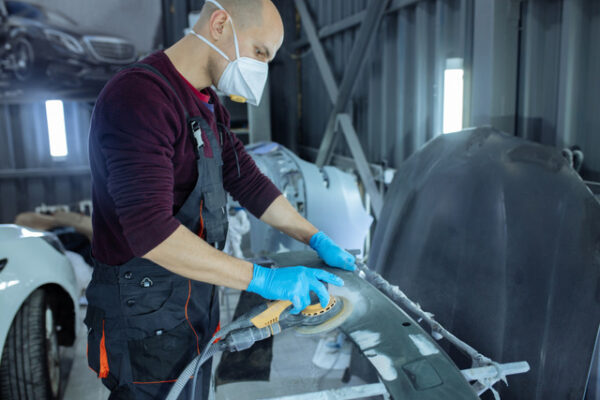 Technicien en carrosserie portant un masque de protection et des gants, utilisant une ponceuse orbitale sur une pièce de véhicule dans un atelier.