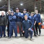 Une équipe dynamique composée de huit employés de Pneus Ratté pose fièrement dans l'atelier mécanique durant la période de changement de pneus. Les membres de la brigade, vêtus de leurs uniformes de travail bleus, affichent un esprit d'équipe renforcé par leur coordination saisonnière.