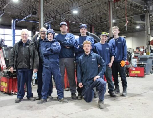 Une équipe dynamique composée de huit employés de Pneus Ratté pose fièrement dans l'atelier mécanique durant la période de changement de pneus. Les membres de la brigade, vêtus de leurs uniformes de travail bleus, affichent un esprit d'équipe renforcé par leur coordination saisonnière.