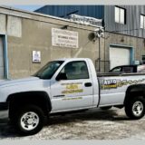 Une camionnette blanche lettrée au nom du Centre de mécanique L.G.M. et arborant le logo Auto Mécano est stationnée sur un sol enneigé devant la façade de l'atelier industriel.