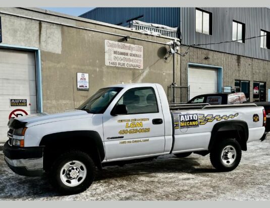 Une camionnette blanche lettrée au nom du Centre de mécanique L.G.M. et arborant le logo Auto Mécano est stationnée sur un sol enneigé devant la façade de l'atelier industriel.