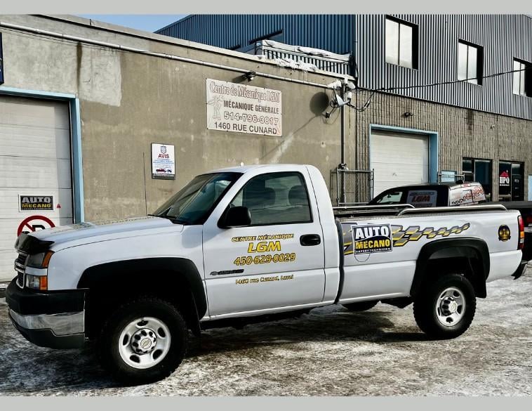Une camionnette blanche lettrée au nom du Centre de mécanique L.G.M. et arborant le logo Auto Mécano est stationnée sur un sol enneigé devant la façade de l'atelier industriel.