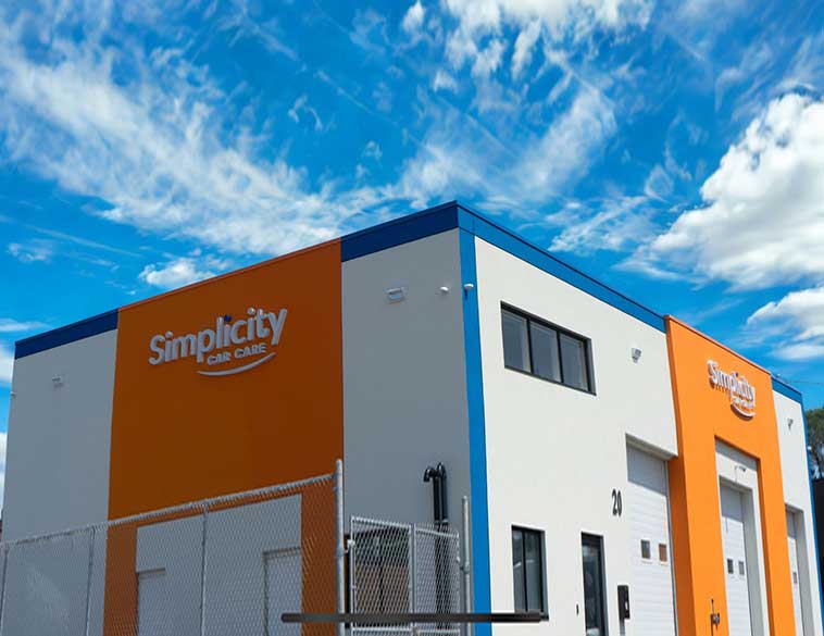 An exterior shot of a modern, multi-colored (orange, grey, blue) building with the text "Simplicity CAR CARE" as a white sign on the façade. The structure has multiple levels and garage doors, all set under a partly cloudy blue sky.