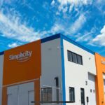 Vue extérieure d’un bâtiment moderne de carrosserie aux couleurs orange, grise et bleue, arborant le logo « Simplicity CAR CARE » sur la façade, sous un ciel bleu parsemé de nuages.