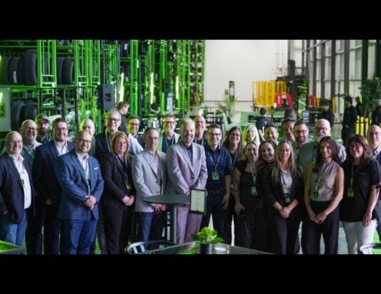 Un grand groupe de personnes souriantes posant pour une photo de groupe à l'intérieur d'un vaste entrepôt, avec de hautes structures de rangement vertes remplies de pneus en arrière-plan.