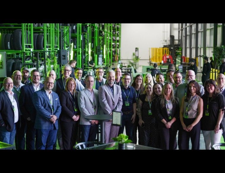 Un grand groupe de personnes souriantes posant pour une photo de groupe à l'intérieur d'un vaste entrepôt, avec de hautes structures de rangement vertes remplies de pneus en arrière-plan.