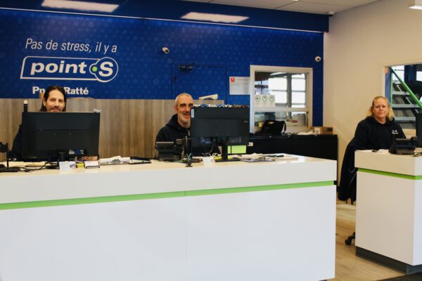 Trois employés souriants de Pneus Ratté, Patrick Abud, Pierre McDuff et Sophie Robitaille, posent derrière le comptoir de service de la succursale d'Anjou sous une affiche bleue Point S.