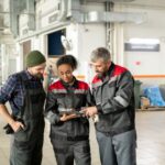 Three auto mechanics standing together in a bright auto repair shop, collaborating and looking at a digital tablet held by a female mechanic.