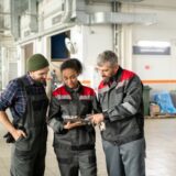 Three auto mechanics standing together in a bright auto repair shop, collaborating and looking at a digital tablet held by a female mechanic.